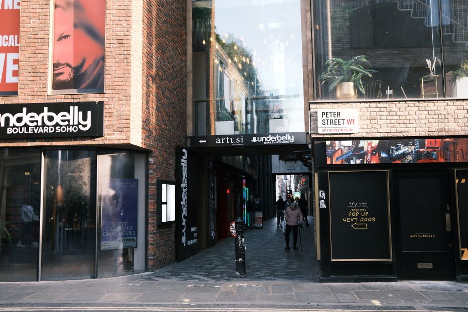 A large decorative teapot-shaped sign with the words 'Antiques Top Arcade' hangs outside a white building on Wardour Street, suspended by black chains. The building features tall windows with white fr